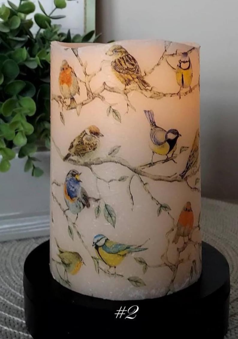 May include: A cylindrical pillar candle with a cream-colored wax surface featuring a printed pattern of various small birds perched on brown branches. The candle is illuminated from within, casting a soft glow, and sits on a black circular base.