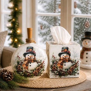 May include: A round glass bottle with a cork stopper and a square tissue box, both featuring a printed illustration of a snowman wearing a plaid scarf and top hat, surrounded by pine cones and greenery, with a white tissue protruding from the box.