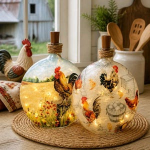 May include: Two decorative glass bottles with rooster and farm scene designs. Each bottle has a wooden stopper and is wrapped with twine. The bottles are lit with small fairy lights, creating a warm glow. They sit on a round woven mat.
