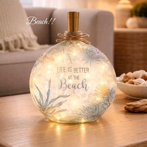 Peut inclure: Bouteille décorative en verre avec bouchon doré, ornée d'un motif sur le thème de la plage. La bouteille est illuminée par de petites lumières et décorée de plantes et de coquillages bleus et blancs. Le texte sur la bouteille dit "LIFE IS BETTER AT THE Beach".