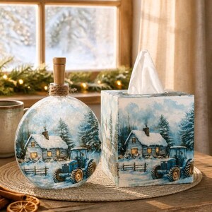 May include: A round glass bottle with a wooden stopper and a square tissue box cover, both featuring a winter scene with a blue tractor, a snow-covered cabin, and pine trees. The items are decorated with a matching blue and white snowy landscape.