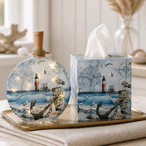 May include: A decorative nautical-themed set featuring a round glass bottle with fairy lights and a tissue box. The bottle and box are adorned with images of lighthouses, anchors, seagulls, and compasses, evoking a coastal theme.