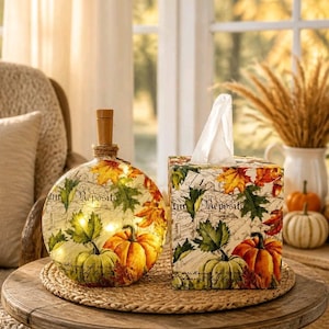 May include: A round glass bottle with a cork stopper and internal lights alongside a square tissue box cover. Both items feature a decorative print of orange and green pumpkins, autumn leaves, and script text on a white background.