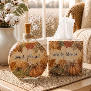 May include: A round glass bottle with a wooden stopper and a square tissue box cover, both featuring a printed autumn design of pumpkins, fall leaves, and the text simply blessed in black script. The items are displayed together as a set.