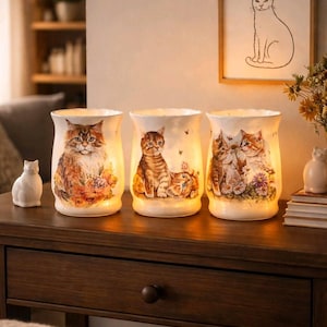 May include: Three decorative cat-themed candle holders with illuminated interiors. Each holder features a different cat illustration with floral accents. A small white cat figurine sits to the left. A framed cat drawing hangs on the wall.