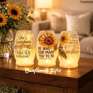 May include: Three illuminated glass candle holders with sunflower designs and inspirational quotes. The text includes phrases like "She's Sunshine" and "Be Who God Made You To Be." The candle holders are on a wooden surface.