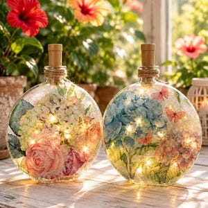 May include: Two round glass bottles with cork stoppers and twine wrapping, each illuminated by internal string lights. The left bottle features pink and white floral patterns, while the right bottle displays blue hydrangea and butterfly motifs.