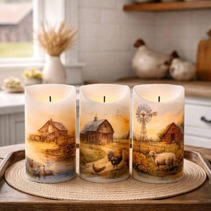 May include: Three white pillar candles with farm scene illustrations. Each candle features a different rural landscape with barns, animals, and a windmill. The candles are displayed on a wooden tray with a woven mat.