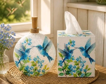 Op de afbeelding: Een ronde glazen fles met houten stop en een vierkante tissuebox, beide versierd met een illustratie van twee blauwe kolibries bij kleine blauwe bloemen en groene bladeren, geplaatst op een rond geweven placemat van jute.