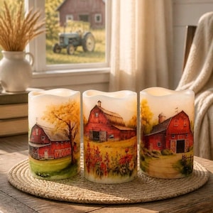 May include: Three cylindrical pillar candles featuring painted scenes of red barns in rural landscapes. Each candle displays a different perspective of a farm building surrounded by trees, fields, and wildflowers, rendered in warm, earthy tones.