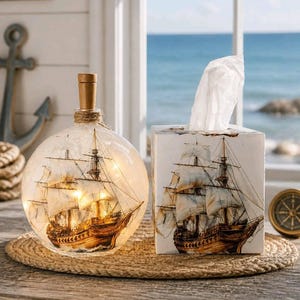 May include: A spherical glass bottle lamp containing a miniature sailing ship with internal warm LED lights, paired with a square white tissue box cover featuring a printed illustration of a sailing ship on the front panel.
