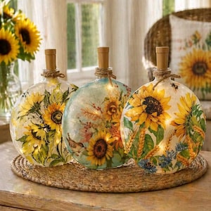 May include: Three decorative glass bottles with sunflower designs and string lights. Each bottle has a wooden stopper and is wrapped with twine. The bottles are placed on a woven mat, creating a warm, inviting ambiance.