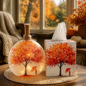 May include: A spherical glass lamp with a wooden stopper and internal lights, paired with a rectangular tissue box. Both items feature a printed design of a tree with vibrant orange and red autumn leaves and a person holding an orange umbrella.