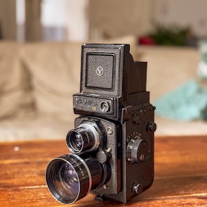 May include: A black Yashica twin-lens reflex camera with a silver lens ring and a top viewfinder. The camera is on a wooden surface. The lens reads "Yashinon". The camera has a classic, vintage design.