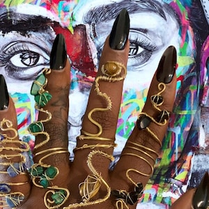 May include: A hand wearing multiple gold wire rings with various gemstones, including green, brown, and black stones. The rings are wrapped around the fingers and some have a spiral design.