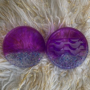 May include: Two round purple and silver glitter resin trays. One tray has three indented sections for holding false eyelashes. The other tray is solid.