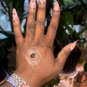 May include: A hand with a copper wire bracelet featuring a central dark stone and a sparkling rhinestone bracelet. The nails are painted light pink with black cross designs.