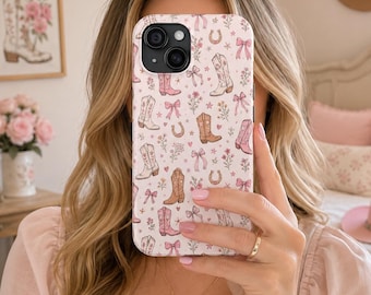 Puede incluir: Una funda de teléfono rosa claro con un patrón repetido de botas de vaquero, lazos, herraduras y flores. La funda tiene un acabado brillante y un recorte para la cámara. Una foto enmarcada de una bota de vaquero está en el fondo.