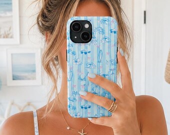 Puede incluir: Funda de teléfono azul claro con un diseño de playa con gafas de sol, tablas de surf y palmeras. La funda tiene rayas verticales y se sostiene frente a una persona. La persona lleva un vestido floral azul y blanco y un collar de estrella de mar.