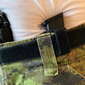 May include: A black handgun with a black holster attached to a black belt. The belt is worn over camouflage pants.