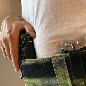 May include: A black handgun in a holster attached to a black belt with a green camouflage pattern. A silver multi-tool is also attached to the belt.