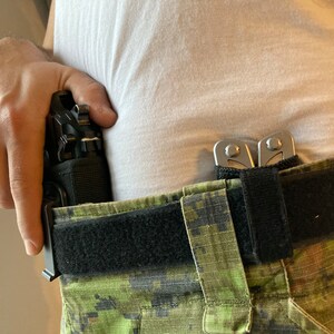 May include: A black gun holster with a black gun inside is attached to a black belt with a green camouflage pattern. A silver multi-tool is attached to the belt.