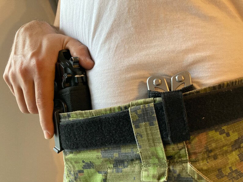 May include: A black gun holster with a black gun inside is attached to a black belt with a green camouflage pattern. A silver multi-tool is attached to the belt.