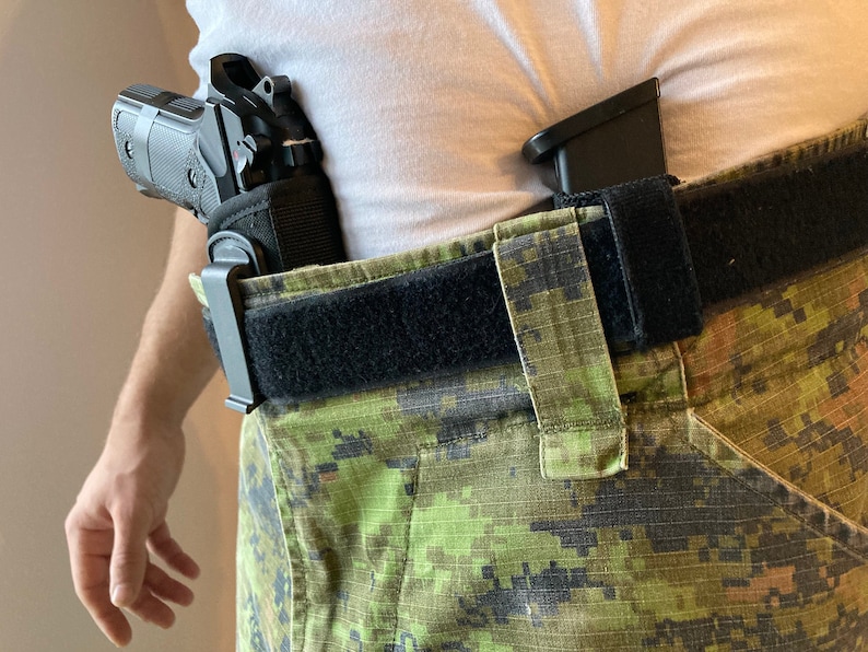 May include: A black handgun with a black holster attached to a black belt. The belt is worn over camouflage pants.