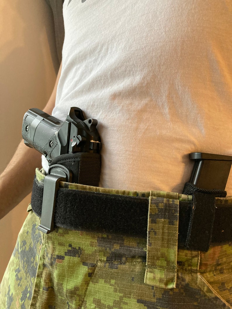 May include: A black handgun with a black holster attached to a black belt with a camouflage pattern. The holster is secured to the belt with a black clip.