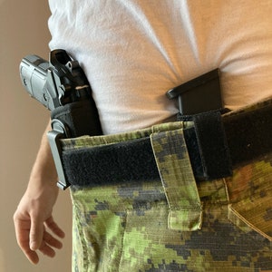 May include: A black handgun with a black holster is worn on a person's waist. The person is wearing a white t-shirt and camouflage pants with a black belt.