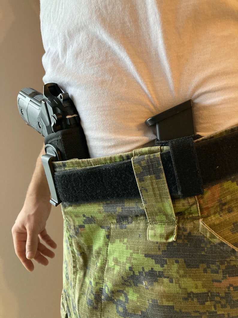 May include: A black handgun with a black holster is worn on a person's waist. The person is wearing a white t-shirt and camouflage pants with a black belt.