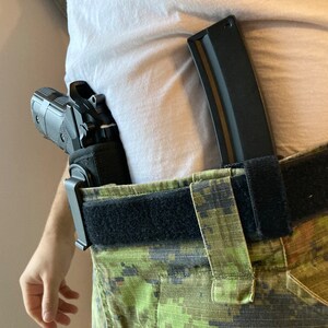 May include: A black handgun in a holster attached to a black belt with a camouflage pattern. A black magazine is attached to the belt.