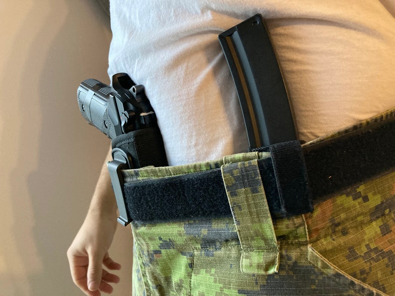 May include: A black handgun in a holster attached to a black belt with a camouflage pattern. A black magazine is attached to the belt.