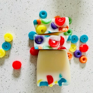May include: A stack of three bars of soap, the top two decorated with colorful, circular shapes. The bottom bar is a solid, creamy color. Scattered around are more of the colorful, circular shapes, suggesting a playful, fun theme.
