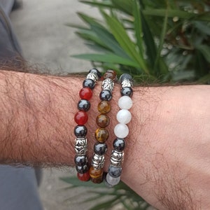 May include: Three beaded bracelets stacked on a wrist. The bracelets are made of red carnelian, tiger's eye, and white moonstone beads. Each bracelet has silver spacer beads with a floral design.