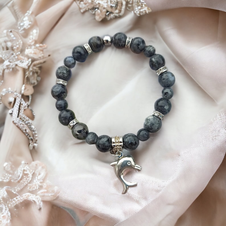 Puede incluir: Un brazalete de piedras grises con un dije de delf&iacute;n plateado. El brazalete est&aacute; hecho de cuentas redondas con detalles plateados.
