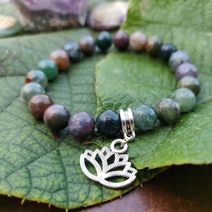 Könnte beinhalten: Ein Armband aus grünem, braunem und violettem Achat mit einem silbernen Lotus-Anhänger. Das Armband liegt auf einem grünen Blatt.