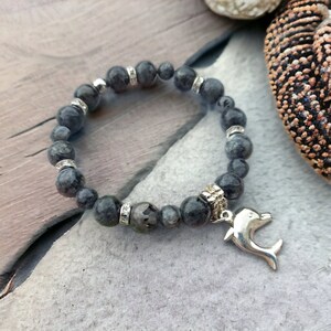 Puede incluir: Un brazalete de piedras grises con detalles plateados y un dije de delf&iacute;n plateado. El brazalete est&aacute; hecho de cuentas redondas con un patr&oacute;n moteado.