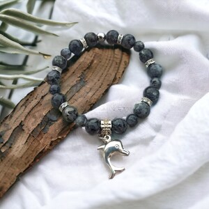 Puede incluir: Un brazalete de piedras preciosas negras y grises con un dije de delf&iacute;n plateado. El brazalete est&aacute; hecho de cuentas redondas y tiene un cierre plateado.