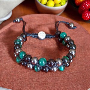Peut inclure: Un bracelet en perles noires et vertes avec un charme argenté. Le bracelet est fait de perles d'onyx noir, de malachite et d'hématite.