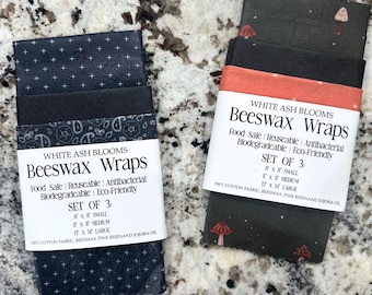 May include: Two sets of White Ash Blooms beeswax wraps. Each set includes three wraps in various sizes: 8x8 inches, 11x11 inches, and 13x14 inches. The wraps are made of cotton fabric, beeswax, pine resin, and jojoba oil. One set has a navy pattern, and the other has a black background with mushroom illustrations.