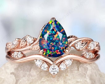 Könnte beinhalten: Ein Verlobungsring mit einem birnenförmigen schwarzen Opal in Roségold, einem zarten Blattdesign und kleinen runden Diamanten. Der Ring liegt auf einem weißen Stein.