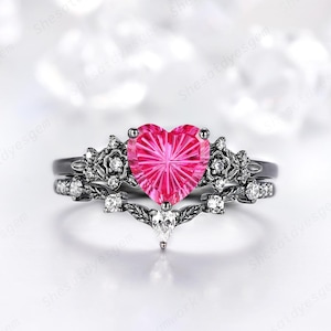 May include: A black metal ring featuring a central heart-shaped pink gemstone with a faceted starburst pattern. The band is designed with intricate floral and vine motifs, accented by small clear round stones and a single pear-shaped stone.