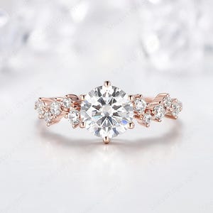 May include: Rose gold engagement ring featuring a large, round, clear gemstone as the centerpiece, surrounded by smaller accent stones. The band has a delicate, floral-inspired design, creating a sparkling and elegant look.