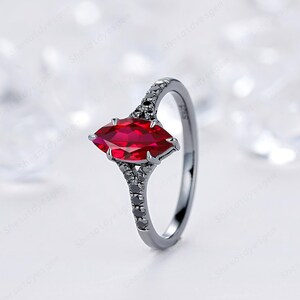 May include: A ring featuring a central marquise-cut red gemstone held by prongs. The band is dark-toned metal and is accented with small, dark, round-cut stones along the shoulders, leading up to the main setting in a split-shank design.