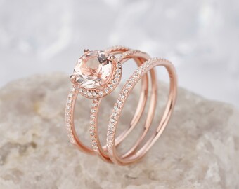 Op de afbeelding: Een verlovingsring van roségoud met een grote, ronde, lichtroze edelsteen omgeven door een halo van kleinere diamanten. De ring wordt geleverd met twee extra roségoud banden, elk bezet met een rij diamanten.