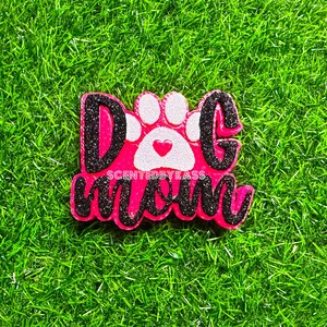 May include: A pink and black decorative item with the words "Dog Mom" and a paw print design. The text and paw print are white and black glitter. The item is on a green grassy surface.