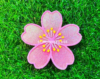May include: A pink, glittery flower-shaped embellishment with yellow stamens. The flower is set against a backdrop of green artificial grass. The text "SCENTEDBYKASS" is visible on the flower.