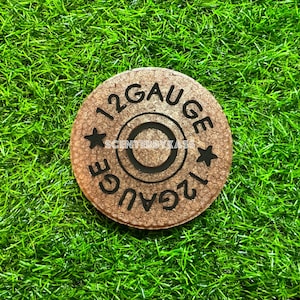 May include: A round, brown coaster with the text "12 GAUGE" and two star designs. The center has a circular design. The text "SCENTEDBYKASS" is also visible. The coaster is set against a background of green artificial grass.