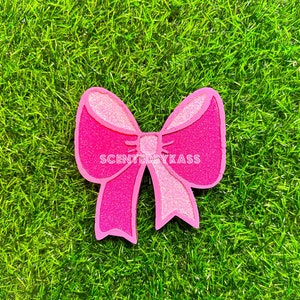 May include: A pink glittery bow-shaped decoration with a light pink center and outline, set against a green grassy background. The text "SCENTEDBYKASS" is printed across the center of the bow.